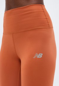Κοντινό πλάνο ενός ατόμου που φοράει αθλητικό κολάν με υψηλή μέση σε καμένη πορτοκαλί απόχρωση, με ανακλαστικό λογότυπο New Balance στο μηρό.