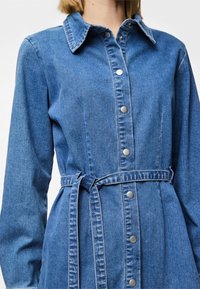 Rochie din denim de culoare albastră medie, cu guler, închidere cu nasturi și o curea din material la talie. Mâneci lungi cu detalii subtile de textură.