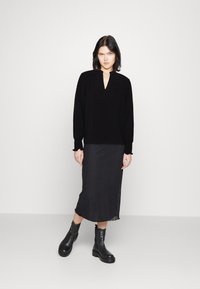 Schwarzes Langarmshirt mit V-Ausschnitt, kombiniert mit einem fließenden schwarzen Rock. Das Modell trägt klobige schwarze Stiefeletten. Glatte Textur, minimales Design.