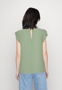 Haut vert à manches courtes avec des accents froncés aux épaules et un détail en trou de serrure au dos, en tissu doux. Porté avec un jean bleu clair.