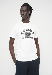 Άσπρο κοντομάνικο t-shirt με μαύρο γραφικό, όπου εμφανίζεται το κείμενο «TOMMY JEANS» και ένας κυκλικός σχεδιασμός. Φοριέται με σκούρο παντελόνι. Ομαλή υφή.