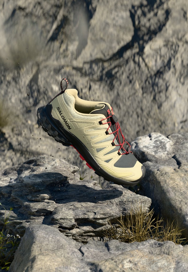 Zapato ligero beige con parte superior de malla transpirable, cordones rojos, suela texturizada y puntera reforzada, diseñado para uso al aire libre.