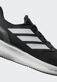Chaussure de sport noire avec des accents synthétiques gris, tige texturée et semelle rembourrée, présentant trois rayures blanches sur le côté et des détails de marque.