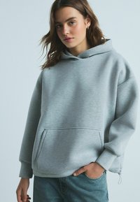 Grå hætte sweatshirt i afslappet pasform, med stor frontlomme og ribbede manchetter. Blød tekstur og minimalistisk design, parret med blå denim jeans.