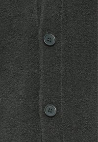 Fekete polár ruhadarab, puha tapintású, két sötét gombbal díszítve. Közeli kép egy gombsorral és enyhe textilfénnyel.