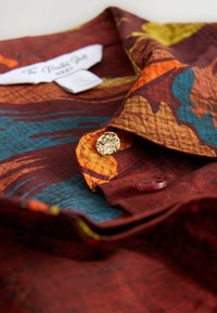 Camisetta a maniche corte di un ricco bordeaux con motivi astratti in arancione, blu e giallo; presenta un colletto classico e bottoni dorati lucidati.