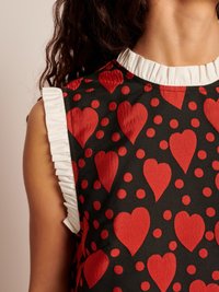 Detalhe de uma blusa preta sem mangas com padrão de corações vermelhos e bolinhas, apresentando um acabamento de babados brancos na gola e nas cavas.