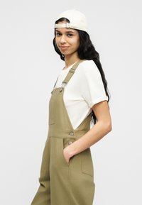 Νεαρός που φοράει μπεζ φορμά, λευκό μπλουζάκι και λευκό καπέλο φοράει ανάποδα, στέκεται πλάγια με το ένα χέρι στην τσέπη, μπροστά σε απλό φόντο.