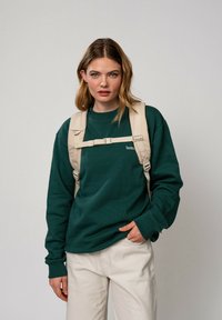 Sweatshirt en coton vert à manches longues, avec un petit logo blanc. Un sac à dos beige est porté sur les épaules. Pantalons de couleur claire.