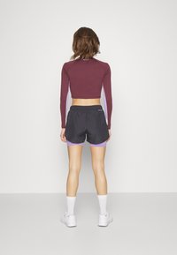 Marjassa pitkähihaine crop top, jossa on violetit sivupaneelit, yhdistettynä mustiin joustavyöhön shortseihin, joissa on violetit reunat. Valkoiset urheilukengät ja sukat.