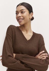 Maglione marrone con scollo a v e maniche lunghe, tessuto testurizzato e polsini a coste. Presenta un delicato dettaglio di cucitura lungo il collo.