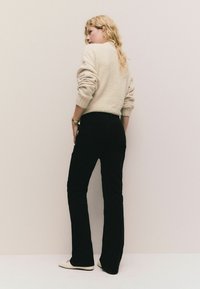 Svetlo béžový sveter s textúrovaným pletením, skombinovaný s čiernymi rozšírenými nohavicami. Modelka má na sebe off-white balerínky a pózuje proti neutrálnemu pozadiu.