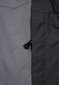 Šedá látková bunda s hladkou texturou, vybavená černou šňůrkou se stahovacím jezdcem a kontrastními švy na pravé straně.