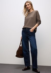 Donna che indossa un maglione marrone e jeans blu scuri, con in mano una borsa in pelle marrone, in piedi contro un muro bianco semplice.