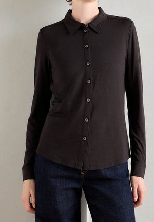 Personne portant une chemise noire ajustée à manches longues boutonnée associée à un jean bleu foncé sur un fond gris clair uni.