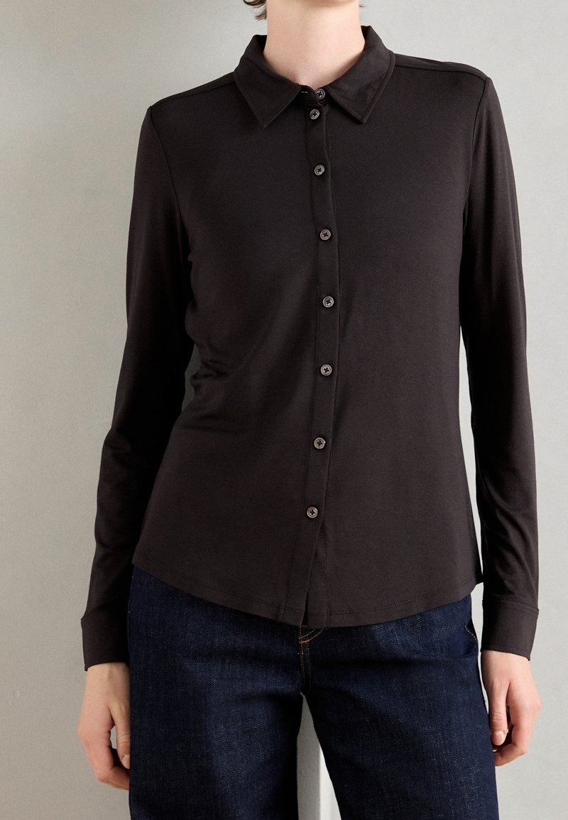 Personne portant une chemise noire ajustée à manches longues boutonnée associée à un jean bleu foncé sur un fond gris clair uni.