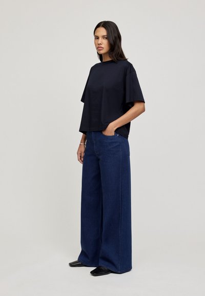 Black cotton t-shirt with wide sleeves paired with high-waisted, loose dark blue denim jeans. Model stands with hands in pockets.