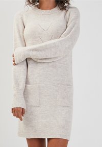 Robe pull tricotée en beige clair, avec un col rond, des poignets côtelés, deux poches plaquées devant, et un motif délicat en losange.