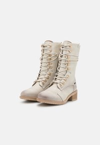 Beige leather ankle boots with a round toe, lace-up design, gold eyelets, and a low stacked heel. Features a textured finish and visible stitching.
