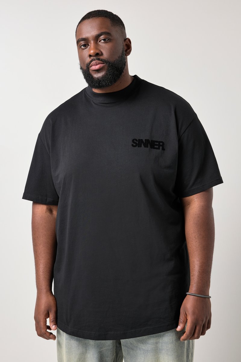 Mann mit Bart, der ein schwarzes T-Shirt mit der Aufschrift "SINNER" auf der Brust, helle Jeans und ein schwarzes Armband trägt, steht vor einem schlichten Hintergrund.