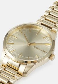 Montre en métal doré avec un cadran rond, un cadran texturé doré, des marqueurs d'heure minimalistes et un bracelet en métal poli. Présente le logo Calvin Klein.
