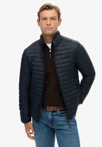Navy puffer jacket with a zip closure, horizontal quilted texture, and a small logo patch on the chest. Paired with a brown sweater and jeans.