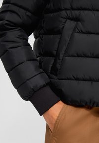 Mano in tasca dei pantaloni marroni, indossando un giubbino trapuntato nero con polsini a coste e dettagli delle tasche anteriori.