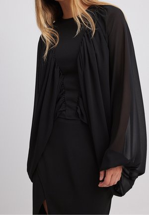 Woman wearing a black dress with sheer, billowy long sleeves and gathered fabric detailing along the front.