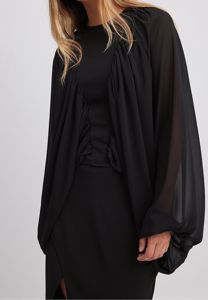 Femme portant une robe noire avec des manches longues transparentes et bouffantes, avec des détails de tissu froncé à l'avant.