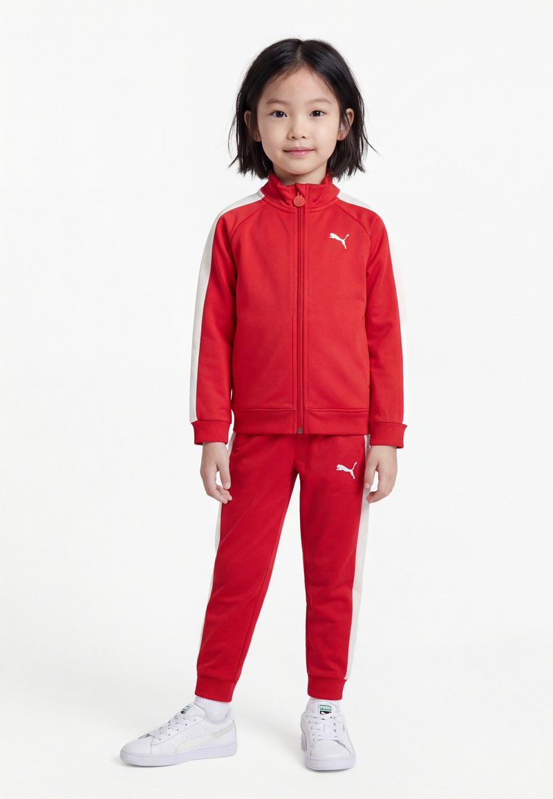 Jeune enfant debout, portant un survêtement rouge Puma avec des bandes latérales blanches et des baskets blanches Puma sur un fond blanc.