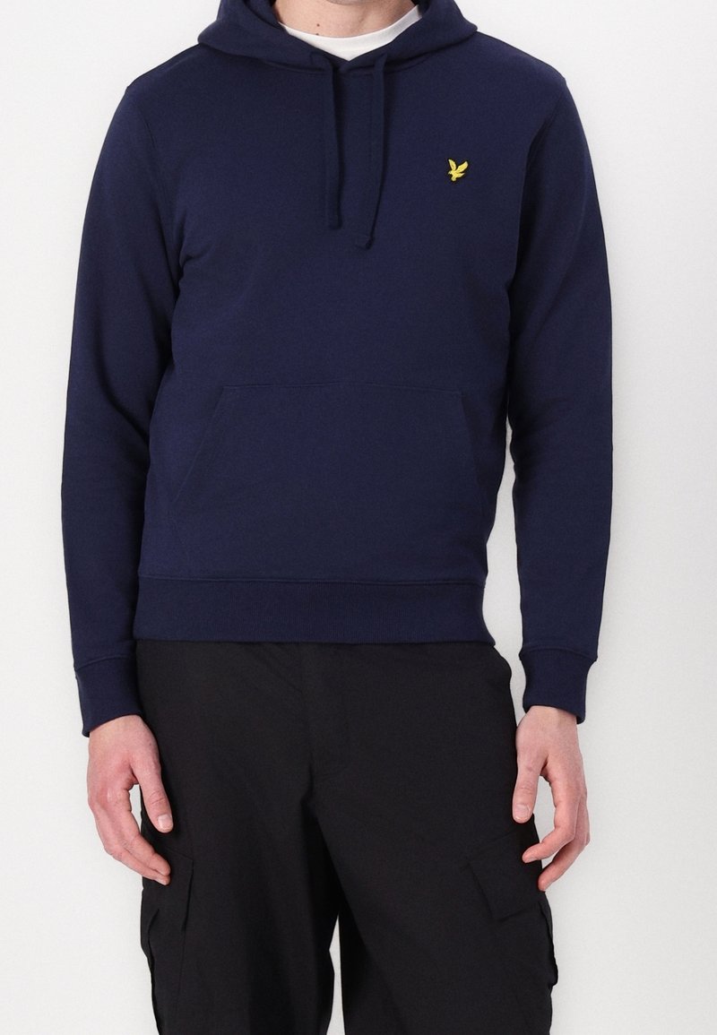 Homme portant un sweat à capuche bleu marine avec un logo d'oiseau jaune sur la poitrine et un pantalon cargo noir, debout sur un fond blanc uni.
