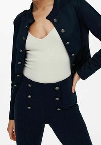 Veste bleu marine et pantalon assorti avec des boutons argentés en accent. La veste est cintrée avec un col rond, et le pantalon est taille haute.