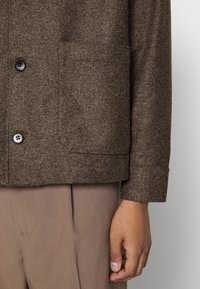 Braune Jacke aus Wollmischung mit einer Tasche, schwarzen Knöpfen und geradem Saum, kombiniert mit hellbrauner Hose. Strukturierter Stoff sichtbar.