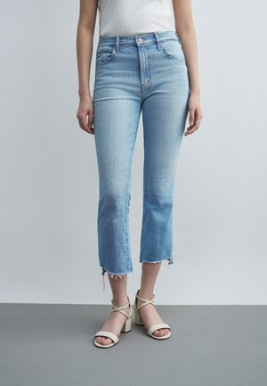 Γυναίκα που φοράει ανοιχτό μπλε κοντά τζιν με ξεφτισμένα τελειώματα και μπεζ πέδιλα με χοντρό τακούνι, στέκεται μπροστά σε απλό φόντο.