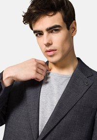 Ciemnoszary żakiet o teksturowanym wykończeniu, z klapami w kształcie notcha i subtelnym akcentem logo; noszony na jasnoszarym t-shircie.