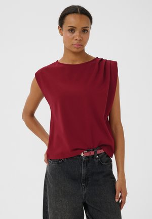 Femme portant un haut sans manches rouge foncé avec des plis aux épaules et un jean foncé taille haute, debout avec une main sur la hanche.