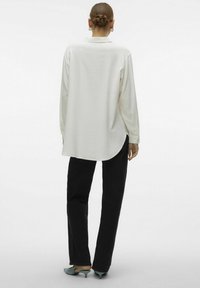 Valkoinen pitkähiha paita, jossa kaulus, väljä istuvuus ja kaareva helma. Yhdistetty mustiin housuihin ja sinisiin kenkiin, minimalistinen muotoilu, sileä pinta.