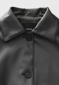 Schwarzer Ledermantel mit strukturiertem Kragen, featuring einer Frontknopfverschluss und einem subtilen Glanz. Das Innenetikett zeigt die Marke "someday."