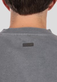 Sivá mikina s rebrovaným golierom, na zadnej strane s čiernou kovovou značkou. Textúra pôsobí mäkko a hladko.