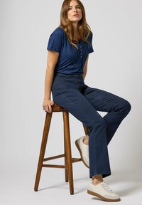 Navy-blaues Kurzarmoberteil mit Knopfdetails, kombiniert mit dunkelblauen Schlaghosen und hellen Sneakers, auf einem Holzschemel vor einem schlichten Hintergrund.