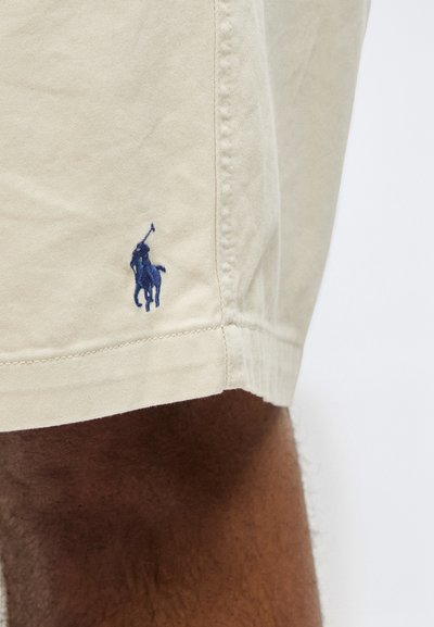 Μπεζ τελείωμα σορτς με μπλε κεντημένο λογότυπο παίκτη πόλο πάνω από το σκούρο δέρμα και τις τρίχες στο μηρό ενός ατόμου.