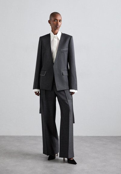 Szürke, szabott blézer kontrasztos zsebkárpittal, széles szárú nadrággal párosítva. Texturált anyag és strukturált fazon, alatta fehér inggel.