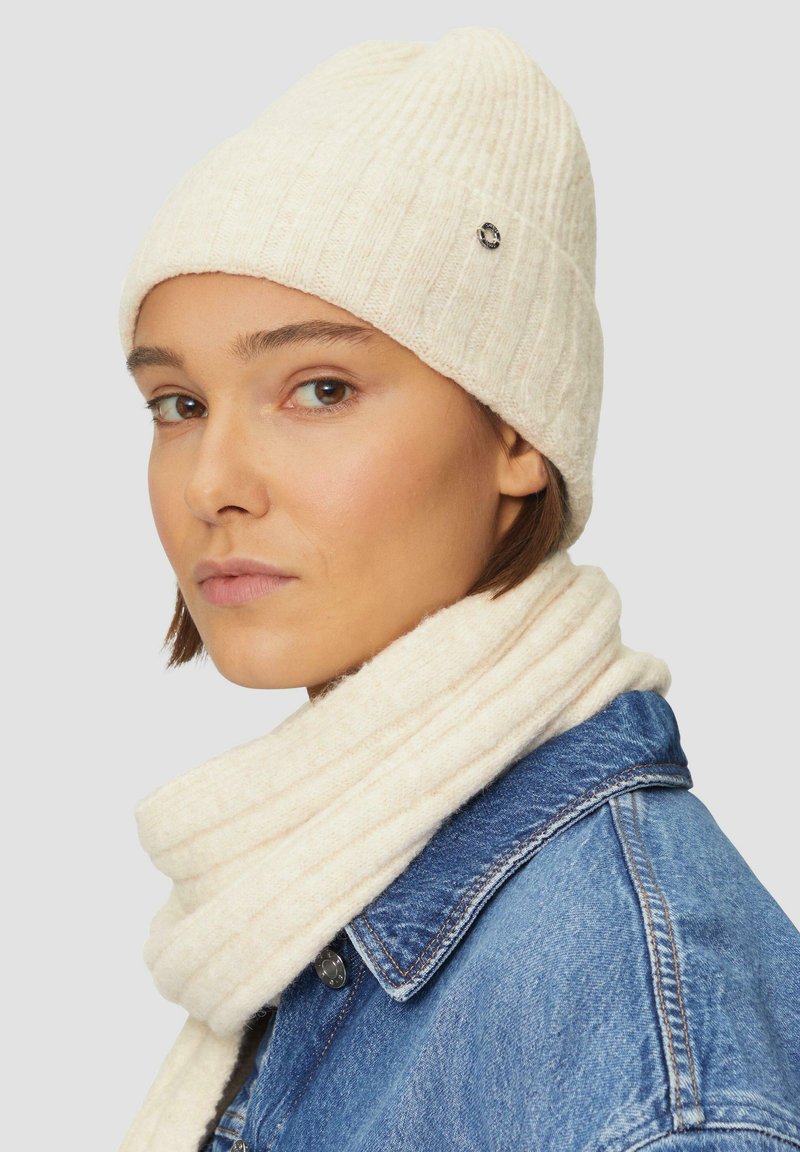Knit cream beanie with ribbed texture and small logo detail, paired with a matching scarf, worn over a denim jacket.
