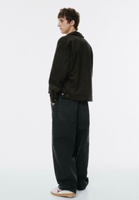 Zwarte suède jas met een losse fit en knoopsluiting bij de mouwen, gecombineerd met zwarte baggy jeans en witte sneakers met rode accenten.