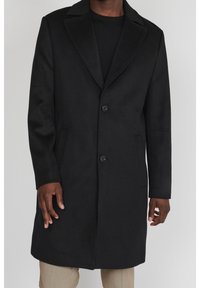 Cappotto di lana nero con due bottoni indossato sopra una camicia nera e pantaloni beige, mostrato su una persona dal collo fino a metà coscia.