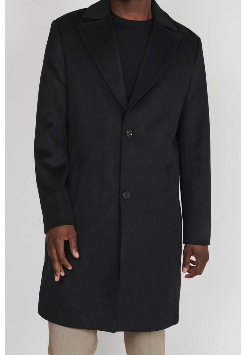 Cappotto di lana nero con due bottoni indossato sopra una camicia nera e pantaloni beige, mostrato su una persona dal collo fino a metà coscia.