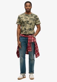 Grøn mønstret t-shirt med korte ærmer, blå jeans og en rød ternet skjorte bundet om taljen. Hvide og sorte sneakers.