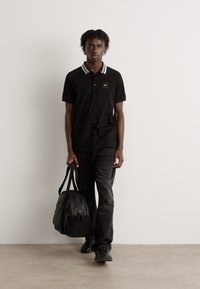 Camisa polo negra con cuello de rayas blancas, combinada con unos jeans negros y zapatillas a juego. Sostiene una bolsa de deporte negra, de pie frente a una pared lisa.