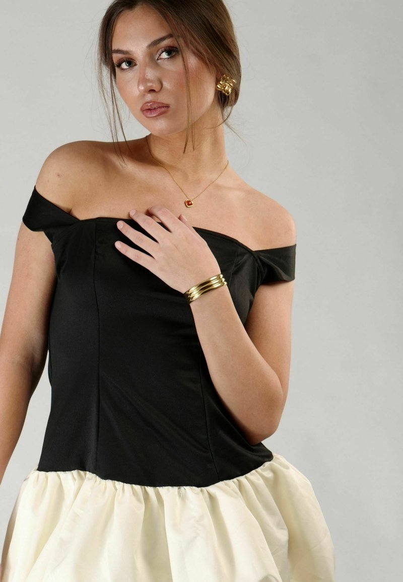 Top nero senza spalline con busto aderente e gonna a balze color crema, adornata con gioielli dorati e una collana a forma di cuore.