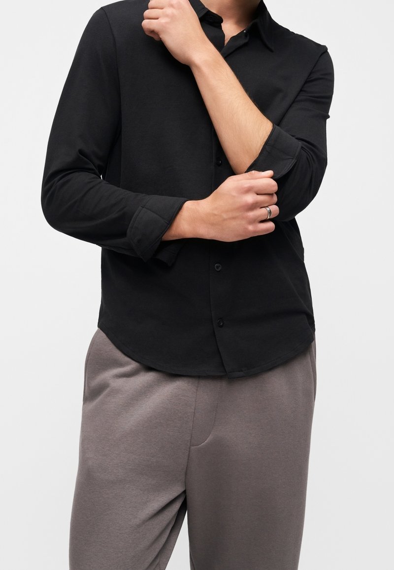 Personne portant une chemise noire boutonnée avec les manches retroussées, associée à un pantalon gris ample, visible de la moitié du torse jusqu’aux cuisses.